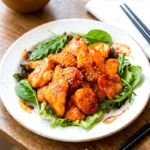 Honey gochujang chicken on a white plate with sesame seeds and green salad