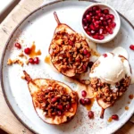 Baked pears with walnut oatmeal crumble served on a plate with melty ice cream, pomegranate arils, and sauce
