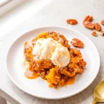 Pumpkin pecan cobbler with vanilla ice cream, caramel drizzle, and pecans on a white plate with a gold spoon.
