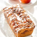 Sliceable apple fritter bread loaf on a white platter with glazed top and two apples in the background