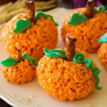 Orange Pumpkin Rice Krispie Treats with pretzel stems and green icing leaves on a cream platter.