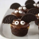 Halloween brownie bite bat with chocolate frosting, cookie wings, and large candy eyes on a white platter.