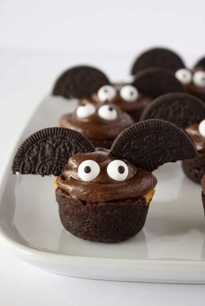 Halloween brownie bite bat with chocolate frosting, cookie wings, and large candy eyes on a white platter.