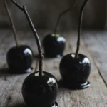Glossy black poison candy apples with tall dark twig-style sticks on a rustic wood board.