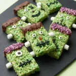 Frankenstein Rice Krispie Treats with chocolate hair, candy eyes, icing faces, and marshmallow bolts on black slate