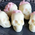 Ivory white chocolate skulls with broken crowns and pale-pink piped cheesecake mousse brains on a dark plate.