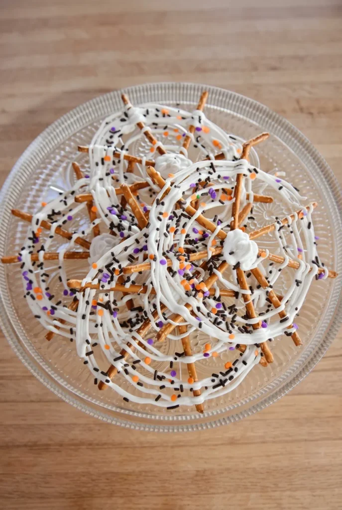 Overlapping Spider Web Pretzels with white candy coating and Halloween sprinkles on a clear glass platter.