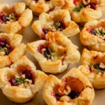 Cranberry Brie bites with flaky puff pastry, melted Brie, cranberry sauce, hazelnuts, and thyme on a beige platter.