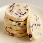 Stacked cranberry orange shortbread cookies with sugar-coated edges, dried cranberries, and orange zest on a cream plate.