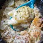 Cheesy scalloped potatoes in a black oval slow cooker with a teal spoon lifting a creamy serving