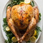 Whole herb butter roast turkey with golden herb-speckled skin, lemon, and fresh herbs on an off-white platter.