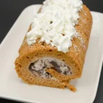 Frozen pumpkin roll with chunky chocolate ice cream swirl, powdered sugar, and whipped cream on an off-white platter.