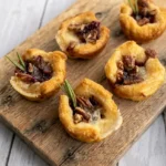 Six baked cranberry Brie bites with ruffled golden puff pastry, cranberry sauce, pecans, and rosemary on a wood board.
