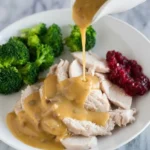 Golden-brown turkey gravy pours from a white gravy boat over sliced roast turkey on a white plate.