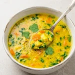 Golden turmeric chicken soup with shredded chicken, carrots, peas, spinach, cilantro, and a spoon in a neutral bowl.