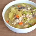White bowl of chunky chicken pot pie soup with shredded chicken, potatoes, mushrooms, carrots, celery, and asparagus.