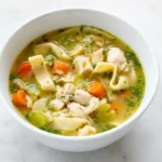 Deep white bowl of brothy chicken noodle soup with wide egg noodles, chicken, carrots, celery, and parsley.