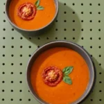 Creamy roasted garlic tomato soup in dark-rimmed bowls topped with roasted tomato rounds and fresh basil