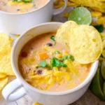 Creamy chicken tortilla soup in a white handled mug with chicken, black beans, corn, cheese, cilantro, and tortilla chips.