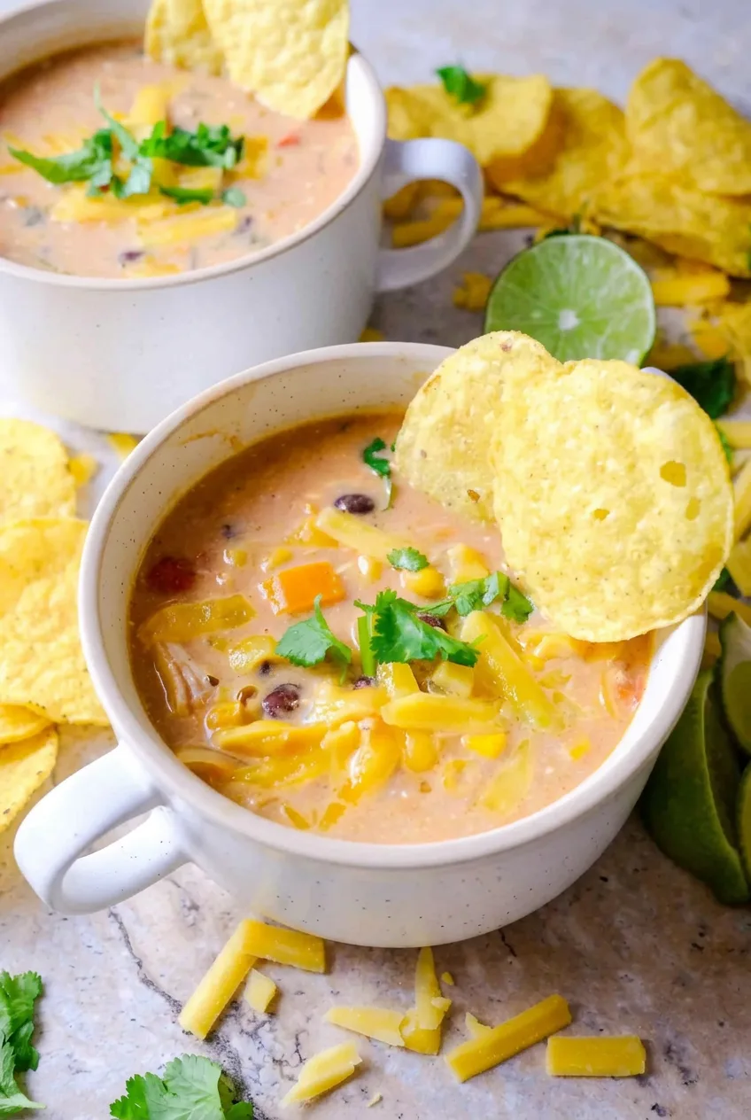 Creamy chicken tortilla soup in a white handled mug with chicken, black beans, corn, cheese, cilantro, and tortilla chips.