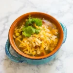 Brothy chicken taco soup with corn, cheddar, crushed tortilla chips, and cilantro in a rustic handled soup mug.
