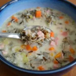 Creamy ham wild rice soup with carrots and celery in a deep blue bowl, with a spoon lifting a chunky bite.