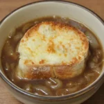 French onion soup in a beige ceramic bowl with onion broth and a centered Gruyère-topped toast.