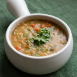Moroccan lentil soup with visible vegetables and cilantro in a white handled bowl on dark green cloth