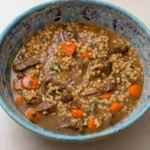 Hearty beef barley soup with beef chunks, pearl barley, carrots, celery, and parsley in a rustic blue-gray bowl.