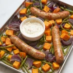 Whole roasted sausages, sweet potatoes, green beans and red onion on a sheet pan with mustard sauce