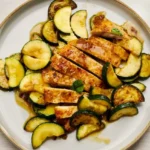 Garlic butter chicken strips layered over zucchini half-moons on a shallow plate with basil leaves