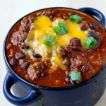 Deep dark bowl of spicy smoky sweet chili with beef, beans, melted cheddar, and green onions