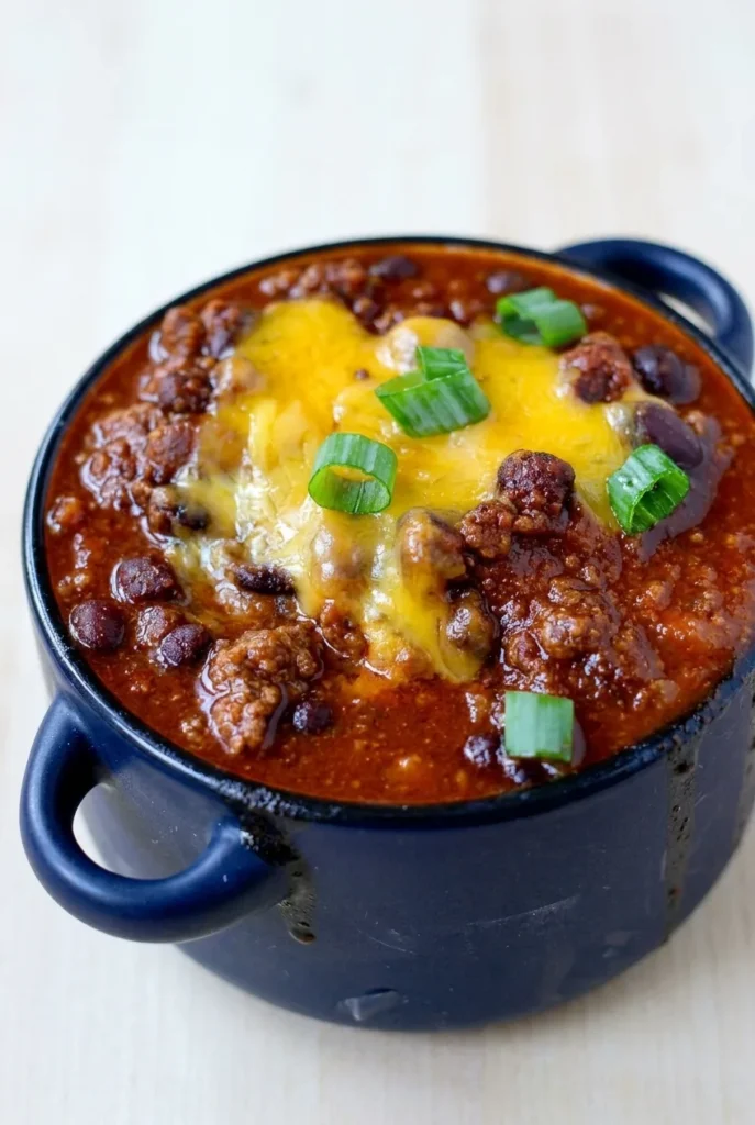Deep dark bowl of spicy smoky sweet chili with beef, beans, melted cheddar, and green onions