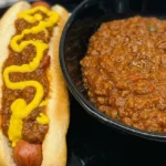 Bowl of thick beef chili beside a chili dog topped with zigzagged yellow mustard