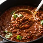 A ladle lifts shredded beef chili with whole pinto beans and cilantro from a dark pot.