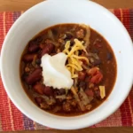 Wide white bowl of saucy ground turkey chili with kidney and black beans, sour cream, and cheddar