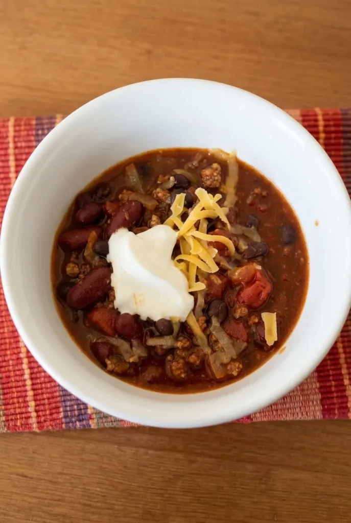 Wide white bowl of saucy ground turkey chili with kidney and black beans, sour cream, and cheddar