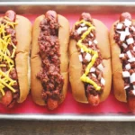 Four hot dog chili dogs on a red-lined tray, finished with cheddar, plain chili, mustard and onion, or onion