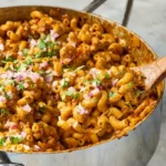Pot of taco mac and cheese with beef, red onion, and cilantro, with a wooden spoon lifting a scoop