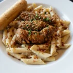Honey pepper chicken strips over creamy bacon penne in a white bowl with parsley and a breadstick