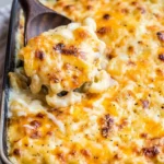 Wooden spoon lifting creamy mac and cheese casserole beneath a spotted golden cheese top