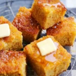 Thick honey butter cornbread squares stacked on a wire rack, showing golden crumb, glossy tops, and butter pats
