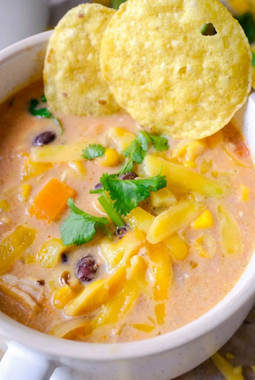 Close view of creamy chicken tortilla soup showing shredded chicken, corn, black beans, melted cheese, cilantro, and a crisp chip.