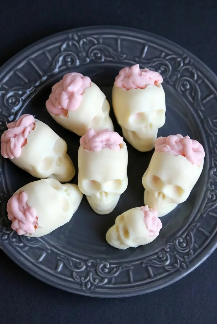 Overhead view of several ivory chocolate skull desserts filled with pink piped brain mousse on a dark serving plate.