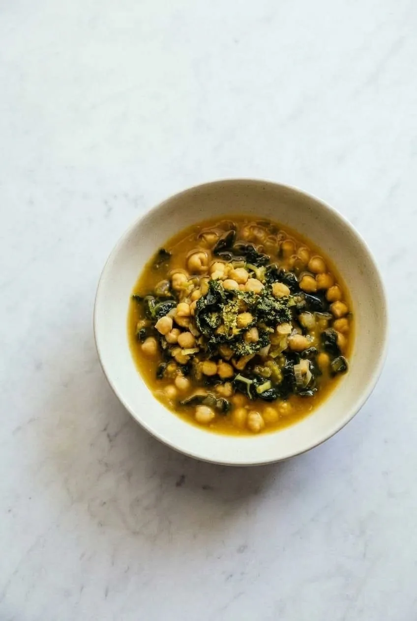 Overhead serving of vegan garlic chickpea soup with kale in a pale bowl on neutral stone