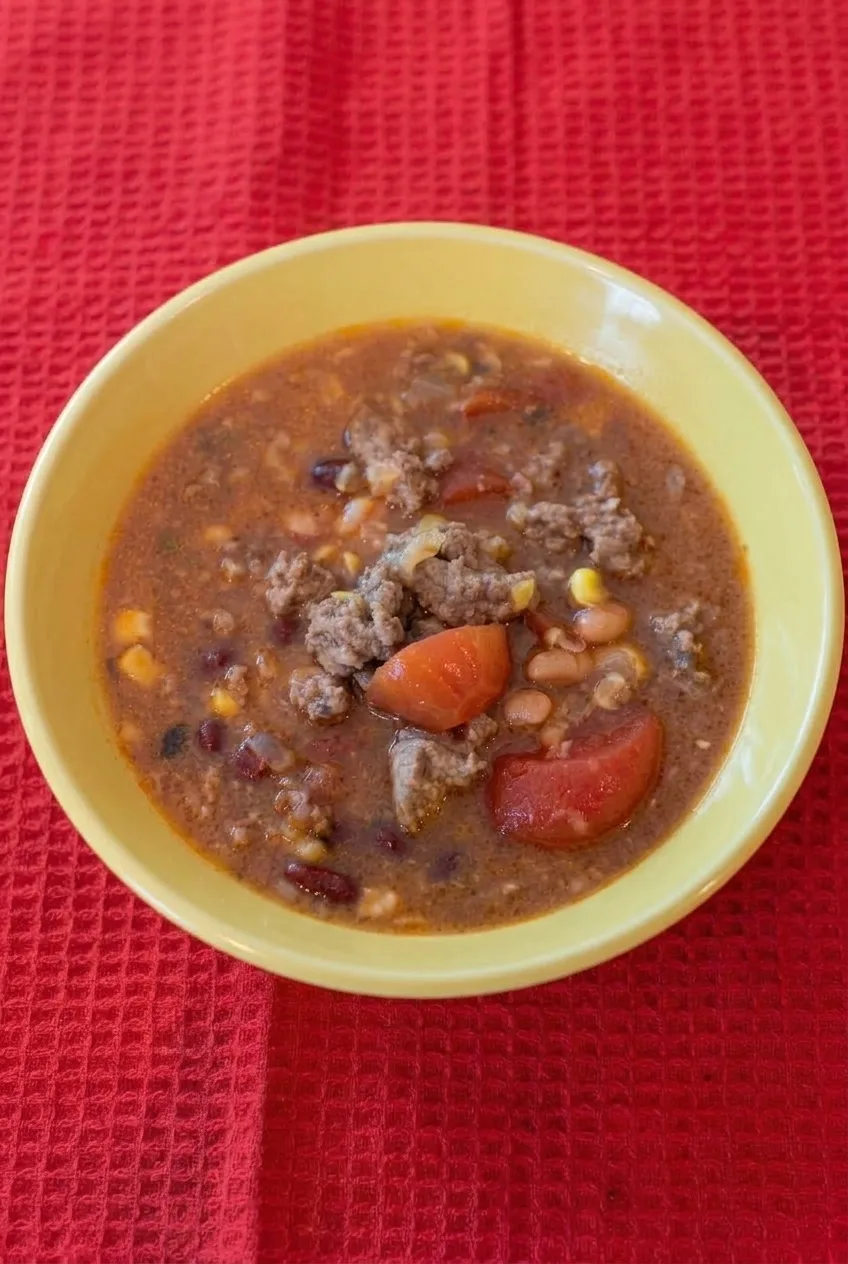 Complete pale yellow bowl of Texas-style beef chili on red textured fabric