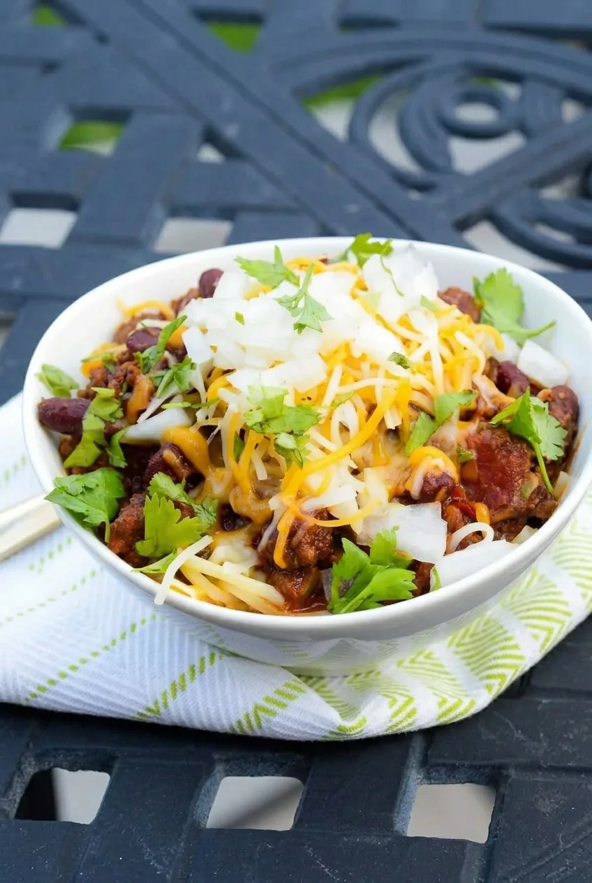 Complete white bowl of Cincinnati chili over spaghetti, with cheddar, onion, and cilantro on a patterned cloth