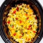 Crockpot ranch chicken with melted cheddar, diced red pepper, and parsley in an oval slow cooker