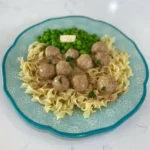 Slow Cooker Swedish Meatballs in creamy gravy over egg noodles, with peas alongside on a turquoise plate