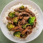 Crockpot beef and broccoli with sesame seeds over white rice in a shallow bowl